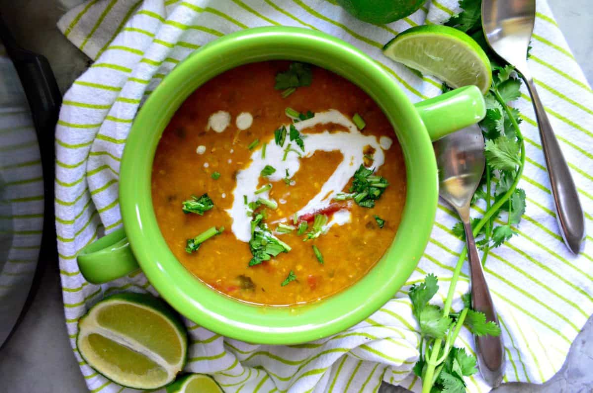 top view of bowl of lentil soup topped with white sauce, and herbs next to spoons and fresh lime.