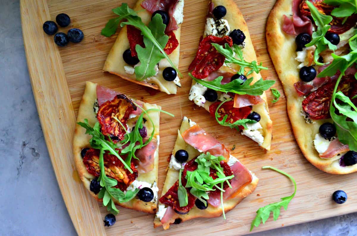 Wood cutting board on a marble countertop with Prosciutto and Blueberry Naan Flatbread cut into pieces.