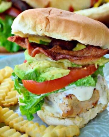 Chicken sandwich with tomato, lettuce, avocado, and bacon with fries on plate.