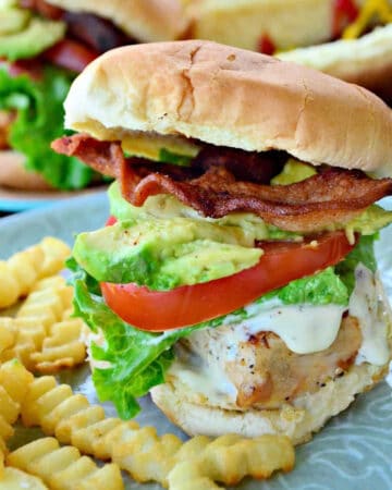 Chicken sandwich with tomato, lettuce, avocado, and bacon with fries on plate.