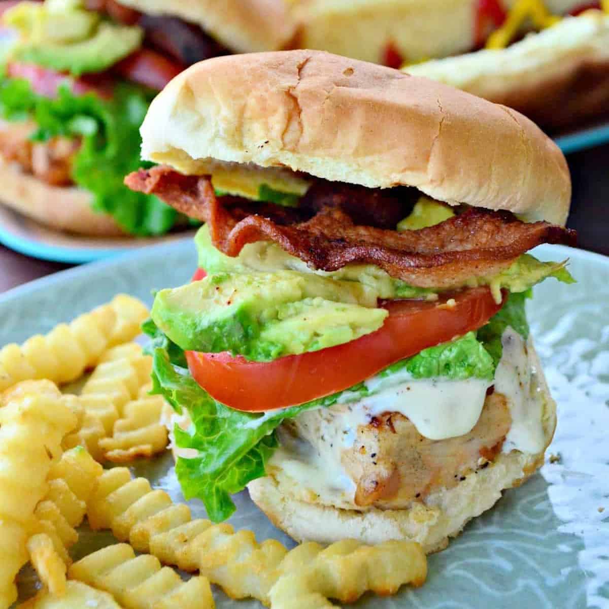 Chicken sandwich with tomato, lettuce, avocado, and bacon with fries on plate.