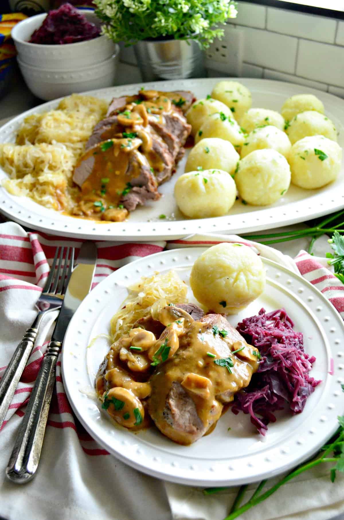 Plated sauerkraut, roasted pork with gravy, and potato dumplings in front of remaining platter by window.