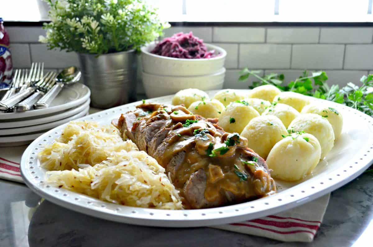Platter of sauerkraut, roasted pork with gravy, and potato dumplings in front of window.