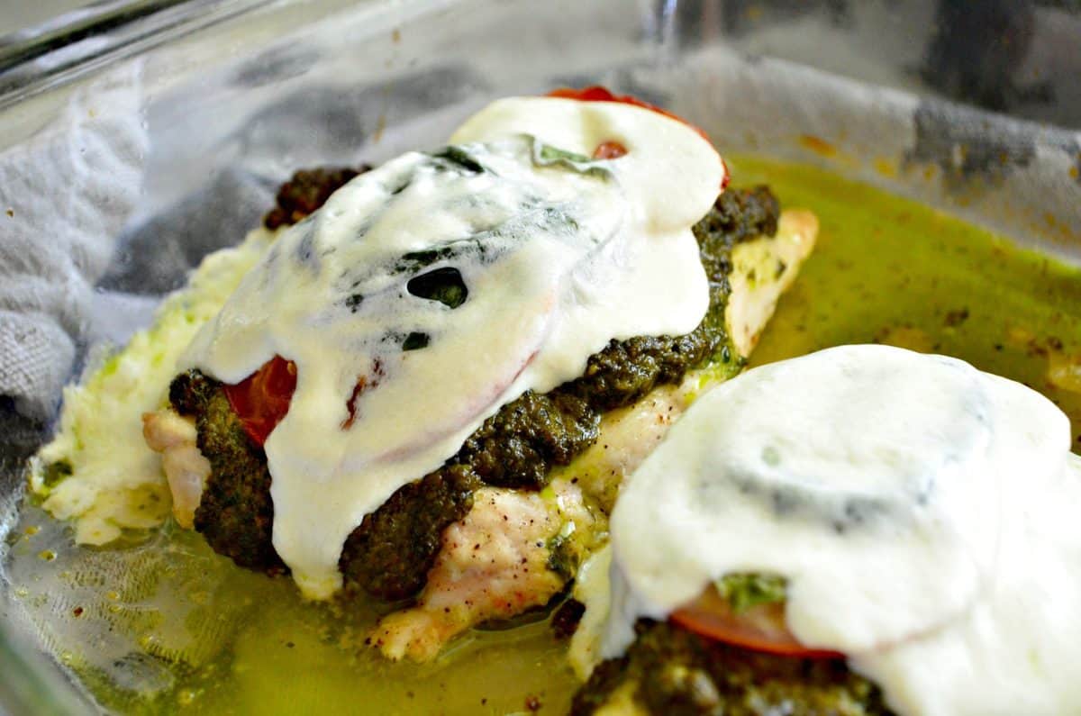 chicken topped with pesto, tomato, mozzarella, and basil in glass baking dish.