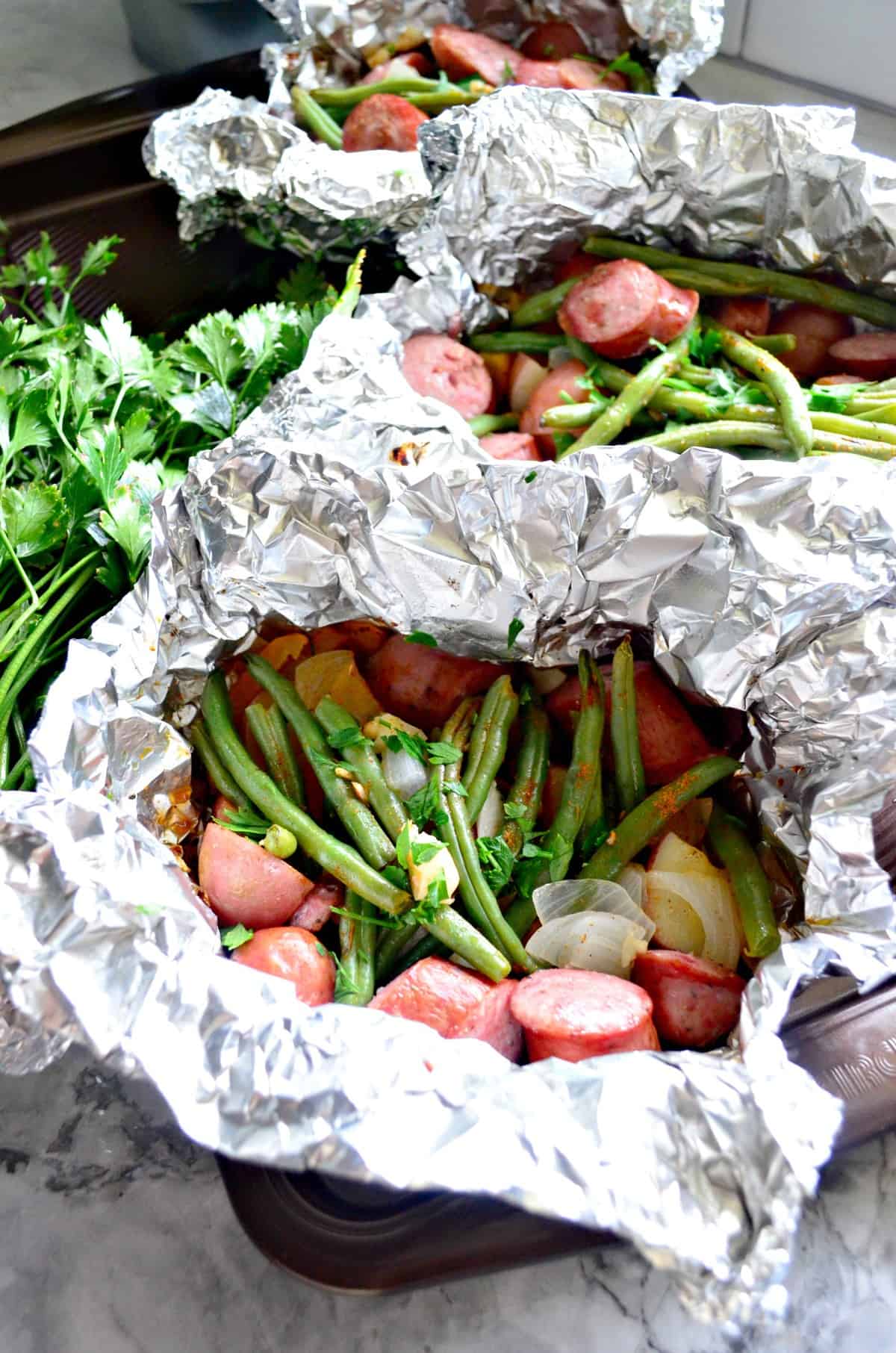 foil packets of kielbasa and green beans next to fresh parsley.