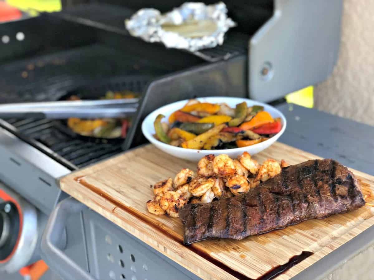 Grilled Skirt Steak next to grilled shrimp and peppers on wooden board next to grill.