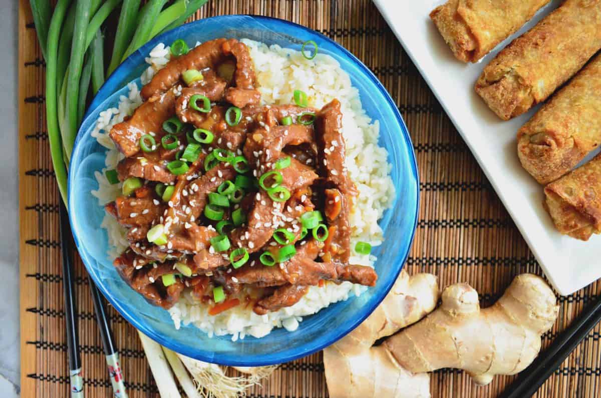 top view strips of mongolian beef with green onions and sesame seeds over bed of rice next to ginger.