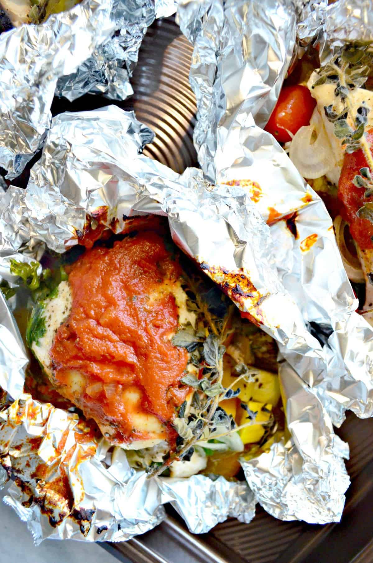 close up of 2 pieces of chicken in an open foil with herbs and red sauce.