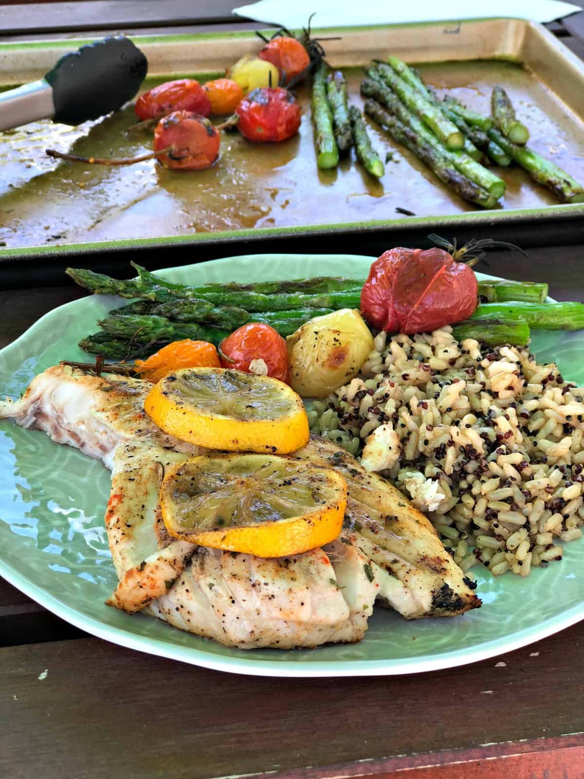 plated meal in front of sheet pan with grilled fish topped with lemon wheels, grape tomatoes, and asparagus.