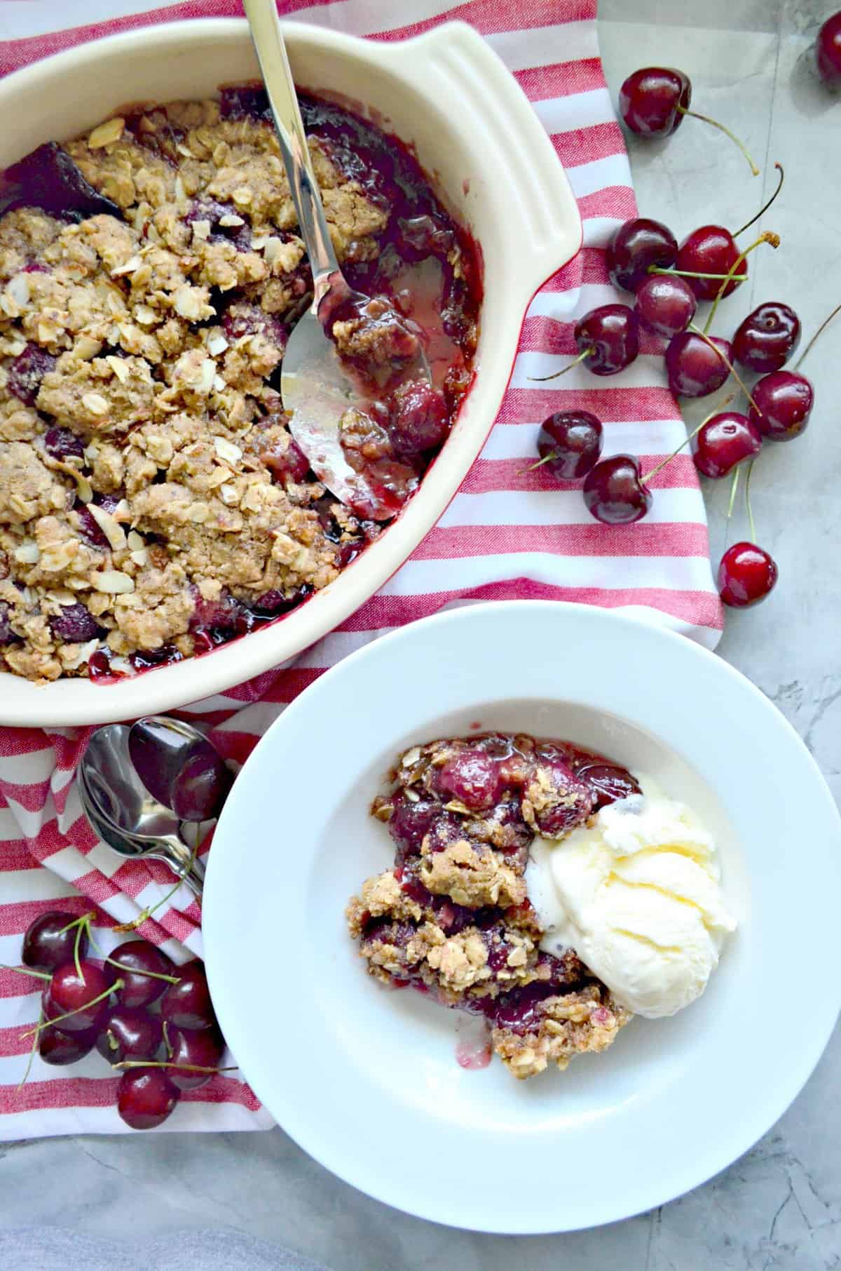 Top view of cherry crisp in a white bowl with ice cream and a baking dish of cherry crisp with spoon.