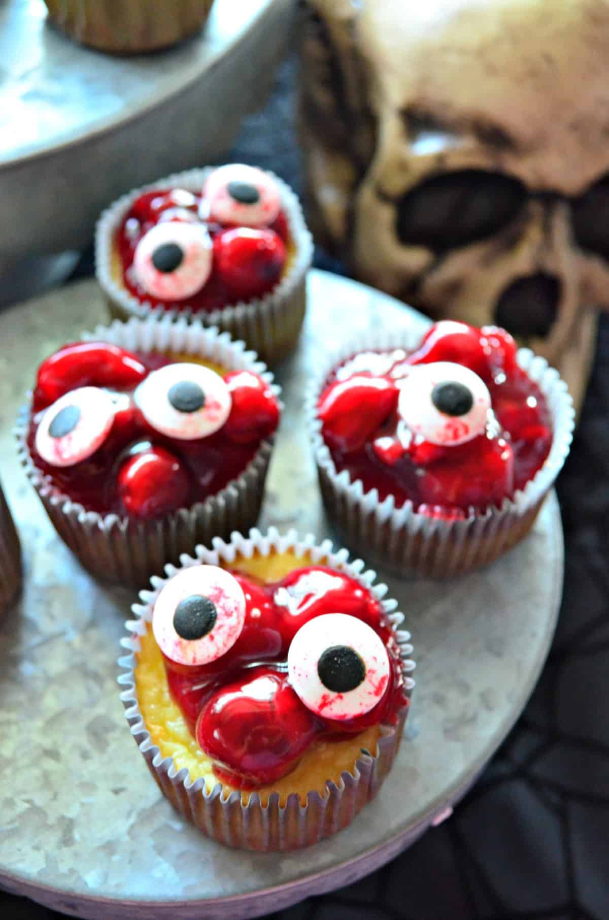 5 cupcakes on cupcake stand topped with red cherries in glaze with candy eyeballs on top.