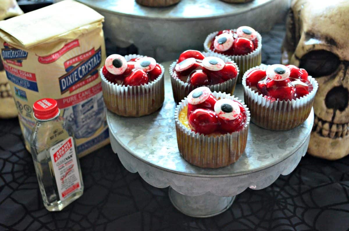 5 cupcakes on cupcake stand topped with red cherries in glaze with candy eyeballs on top next to vanilla extract.