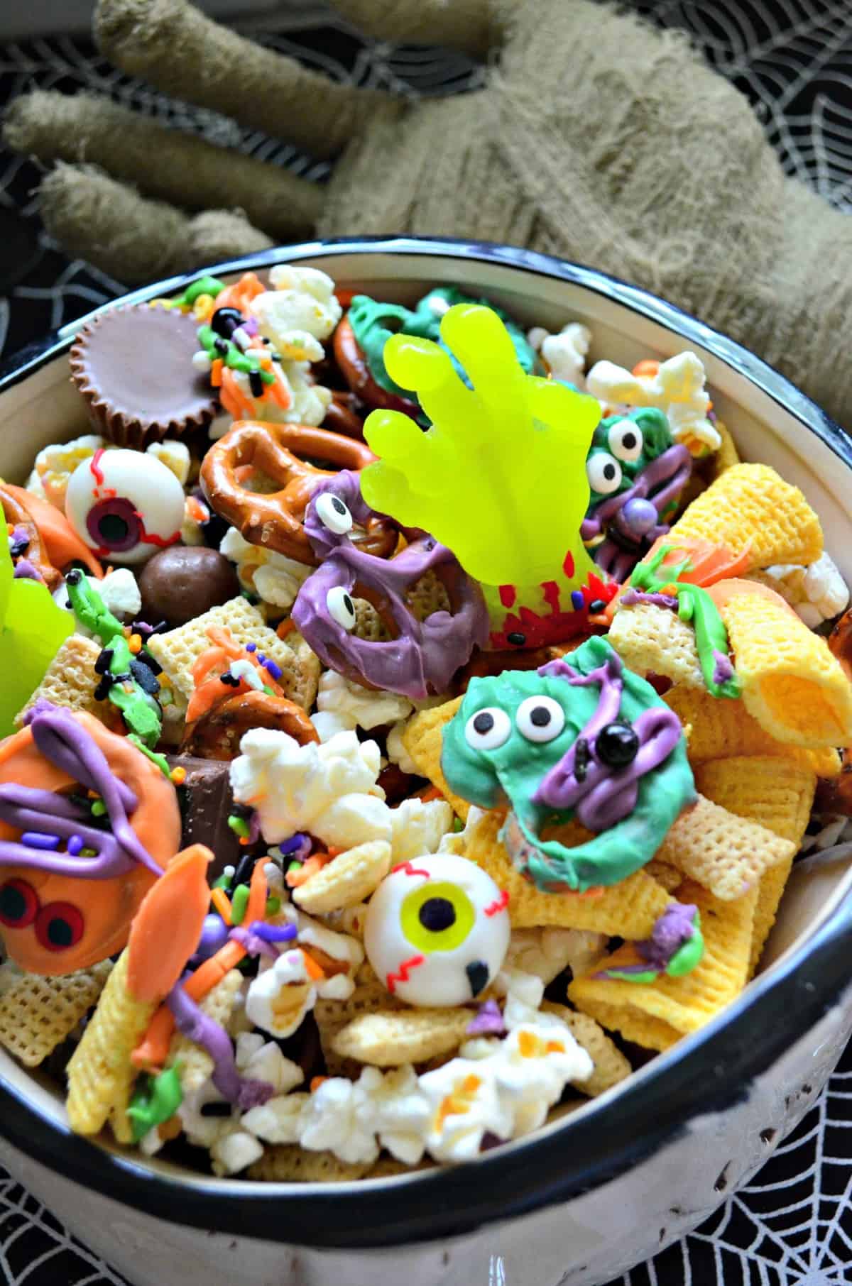 bowl of snack mix with colorful chocolate dipped pretzels and candy eyes with gummy zombie hands.