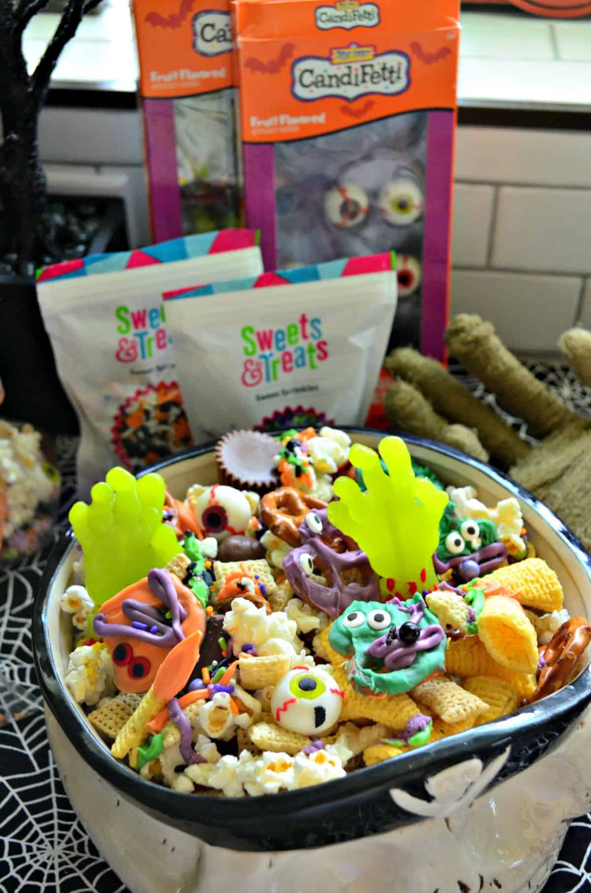 skull bowl of snack mix with colorful chocolate dipped pretzels and candy eyes in front of sweets & treats bags.