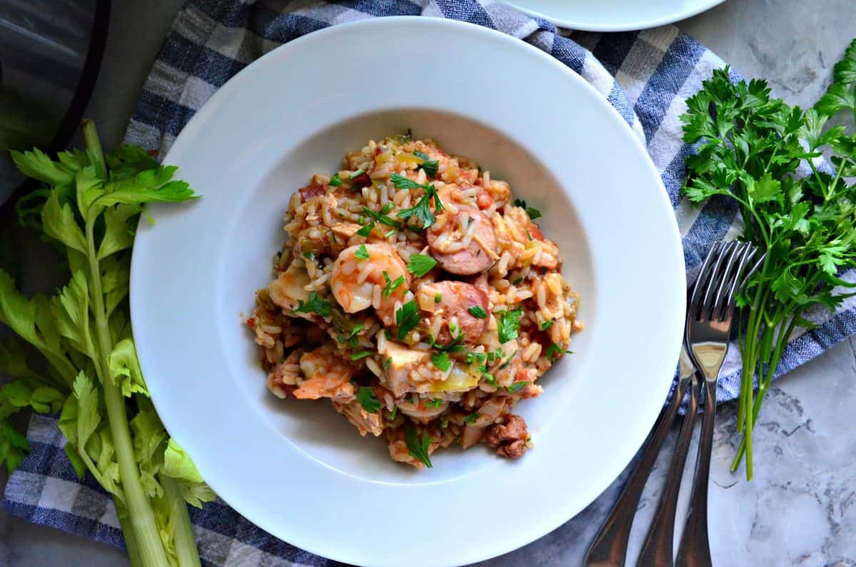 Chicken, Andouille Sausage, and Shrimp Jambalaya in a bowl with rice on tablecloth next to forks and herbs.