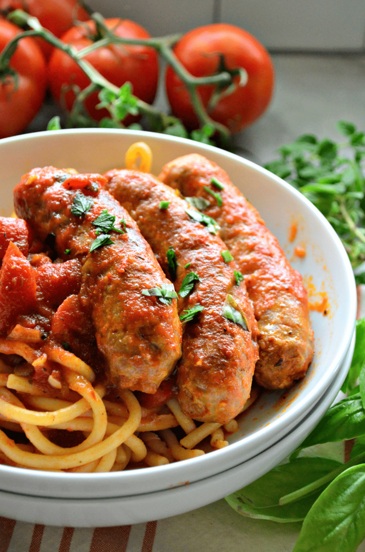 close up bowl of italian sausage with marinara and pasta with vine tomatoes blurred in background.