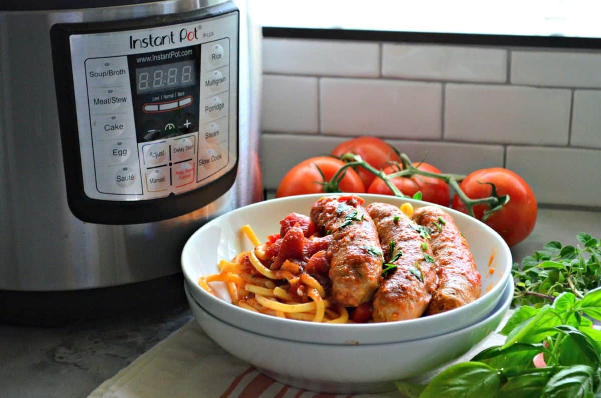 Bowl of italian sausage with marinara and pasta in front of instant pot and tomatoes.