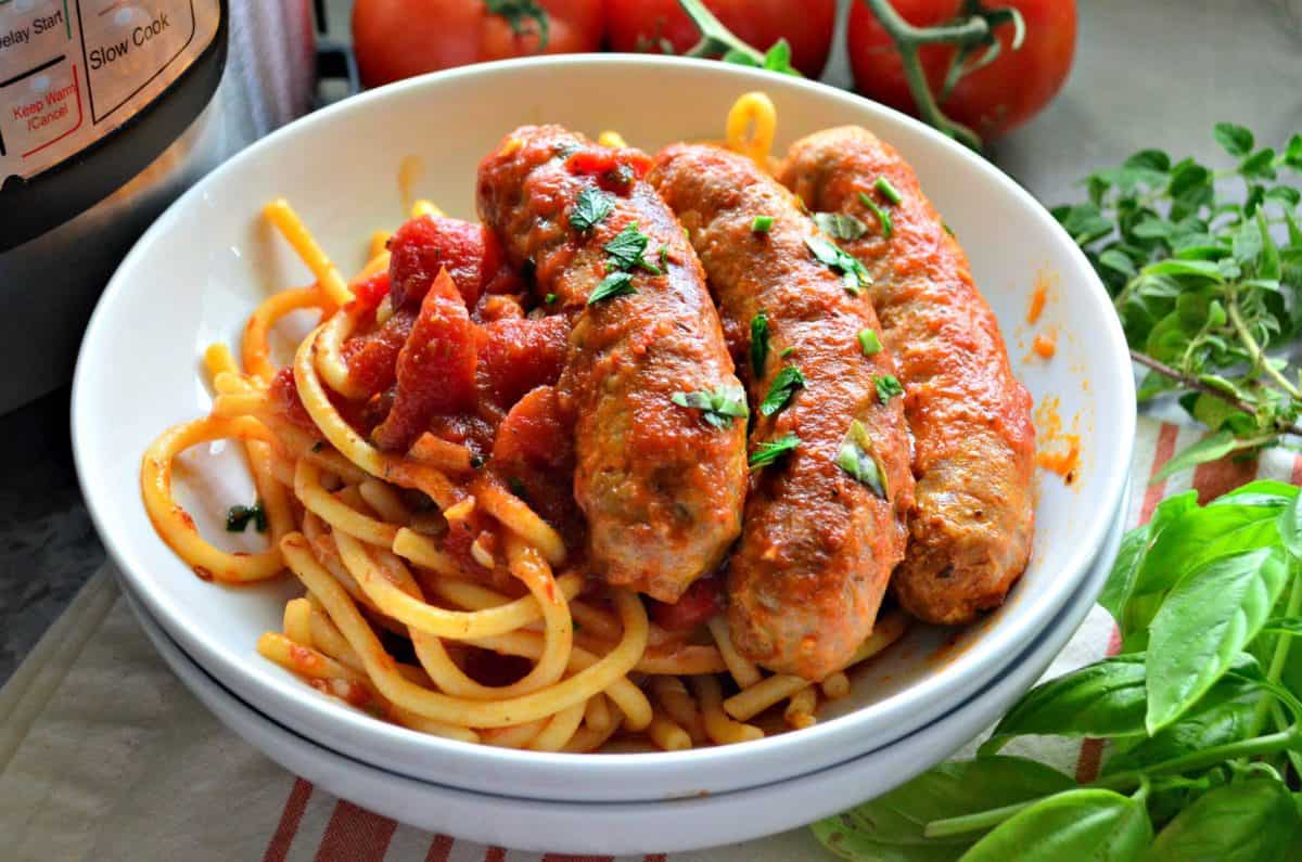 Bowl of italian sausage with marinara and pasta next to oregano and basil.