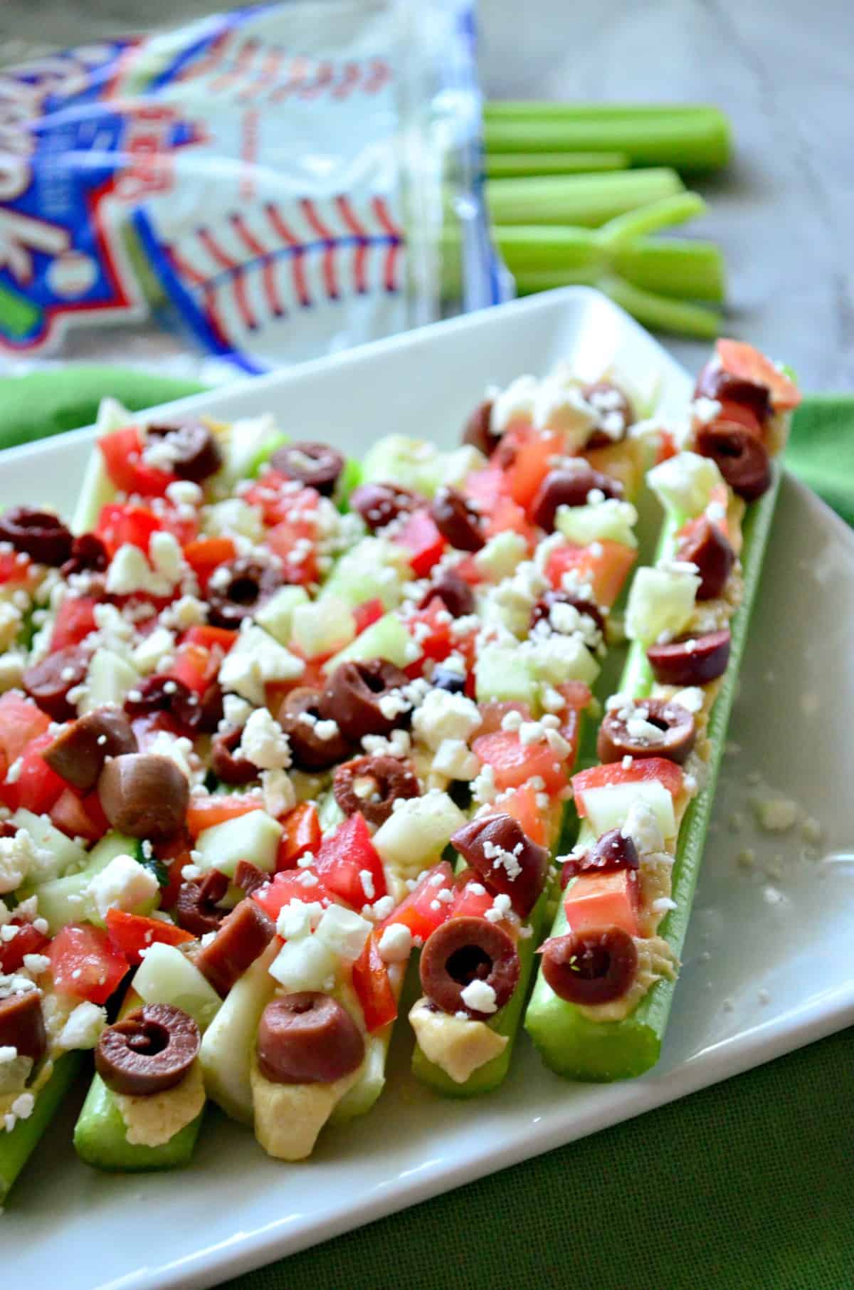 Plated celery sticks stuffed with hummus, feta, kalamata, and tomato with celery blurred in background.