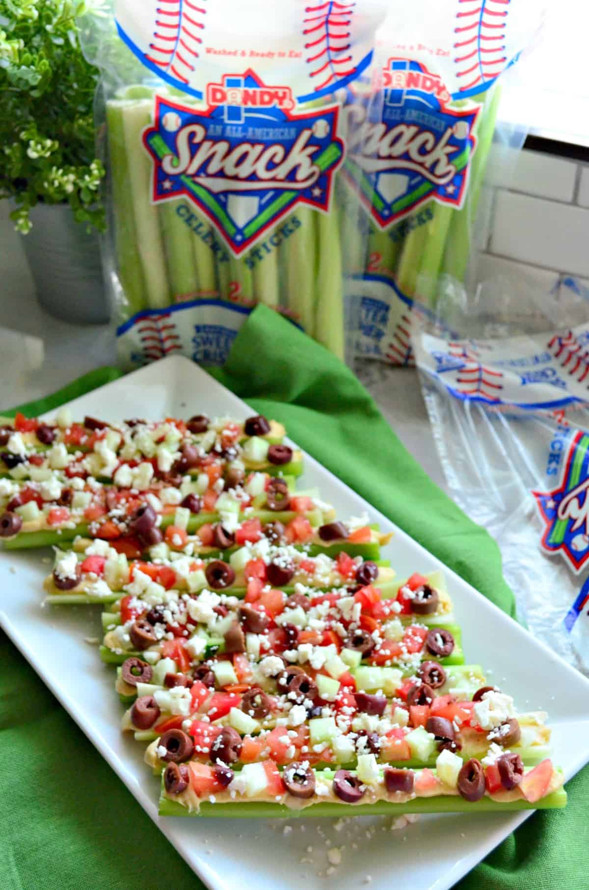 Plated celery sticks stuffed with hummus, feta, kalamata, and tomato in front of fresh celery.