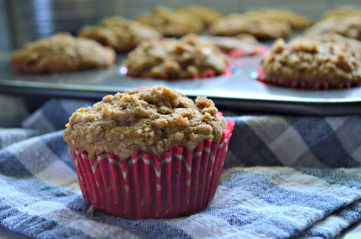 closeup of apple cinnamon streusel muffin in red paper with muffins in muffin tin faded in background.