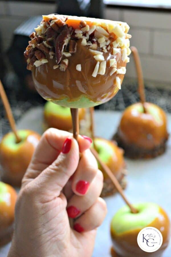 Female hand with red fingernails holding a caramel apple with chopped nuts with logo on right bottom corner.