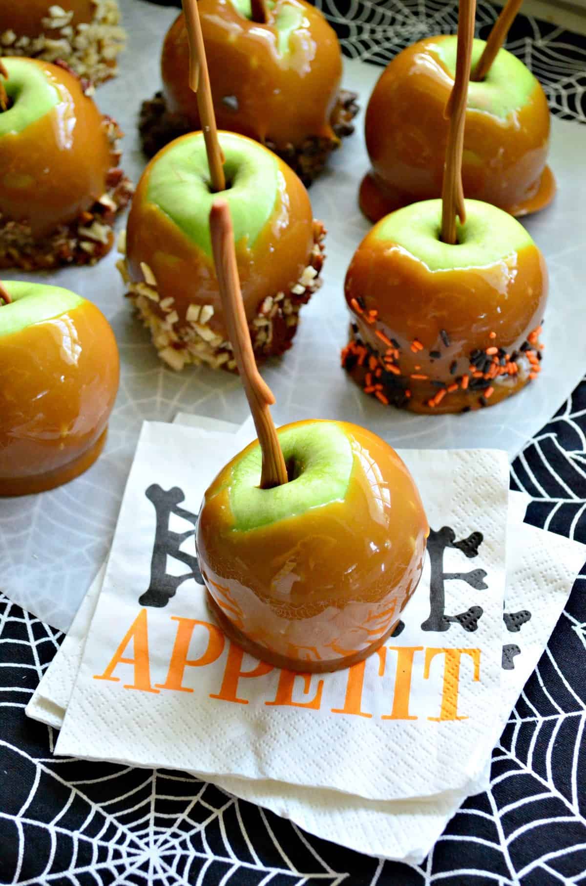 10 green apples dipped in caramel, 5 of which are covered in nuts or sprinkles on spiderweb platter.