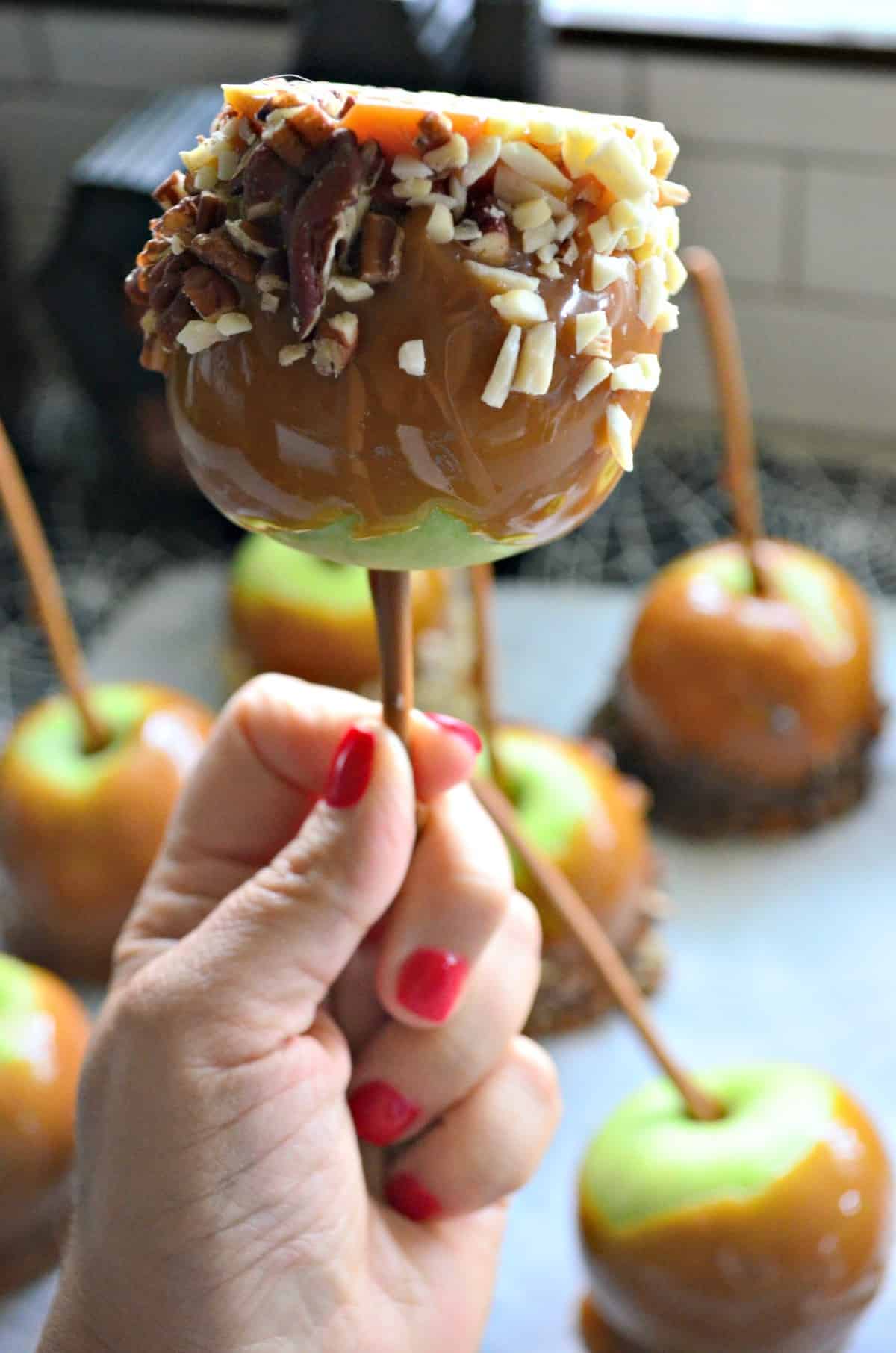 closeup of hand holding up green apple dipped in caramel covered in nuts.