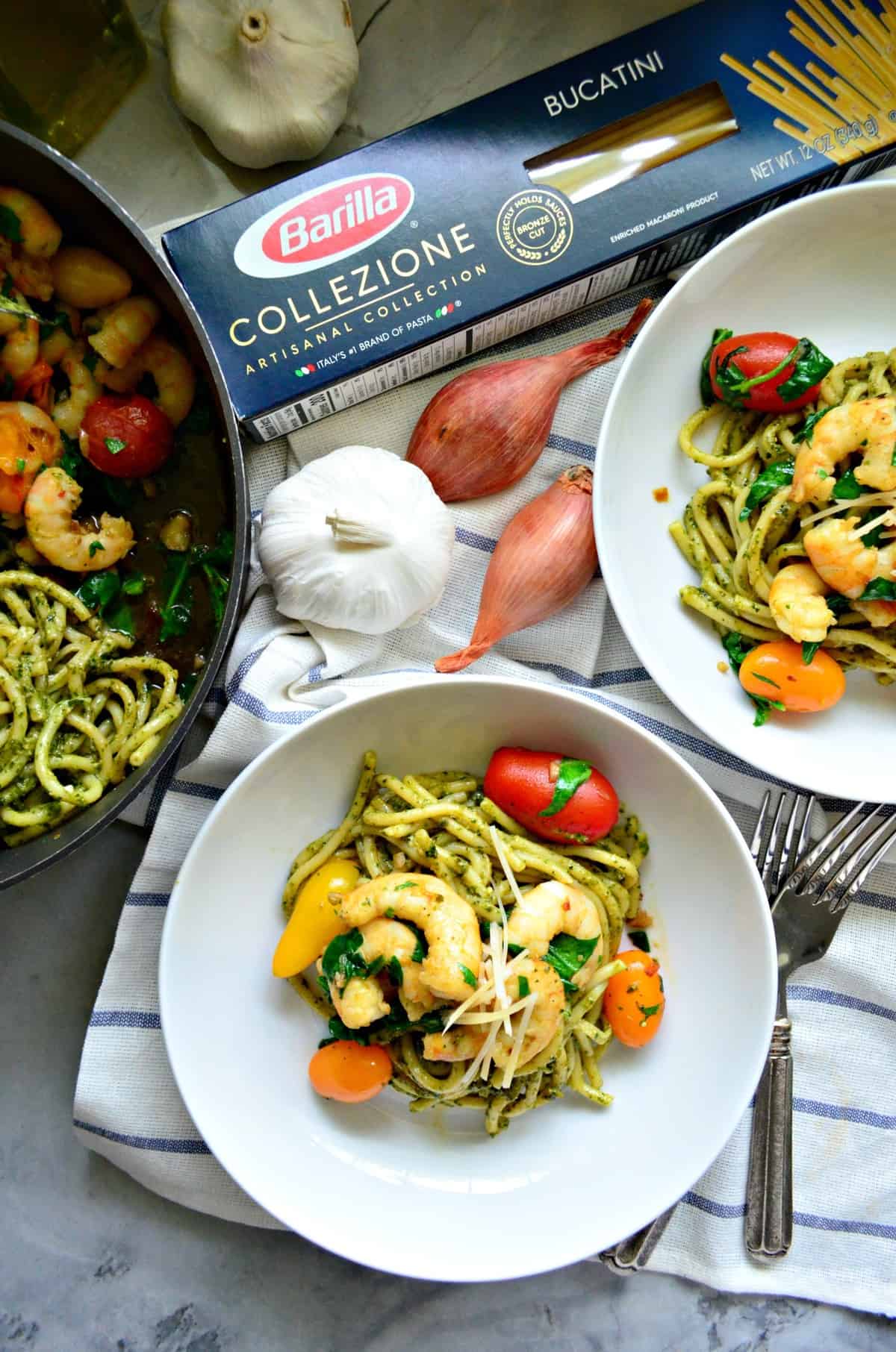 2 bowls of bucatini with pesto, grape tomatoes, shrimp, basil, and cheese on tablecloth with whole ingredients.