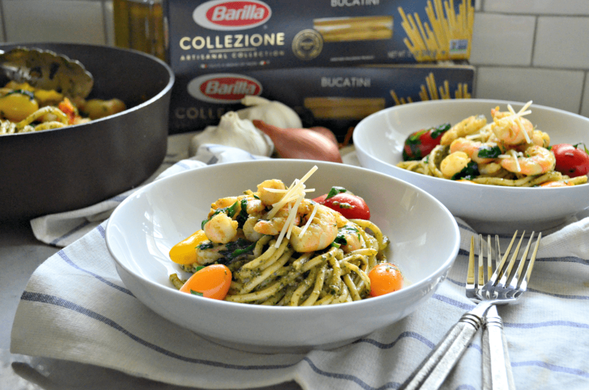 2 bowls of bucatini with pesto, tomatoes, shrimp, basil, and cheese on tablecloth in front of pasta boxes.