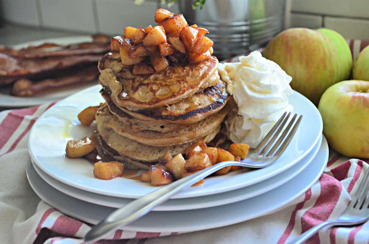 side view stack of plated cinnamon pancakes topped with cubed apples and syrup with whipped cream.