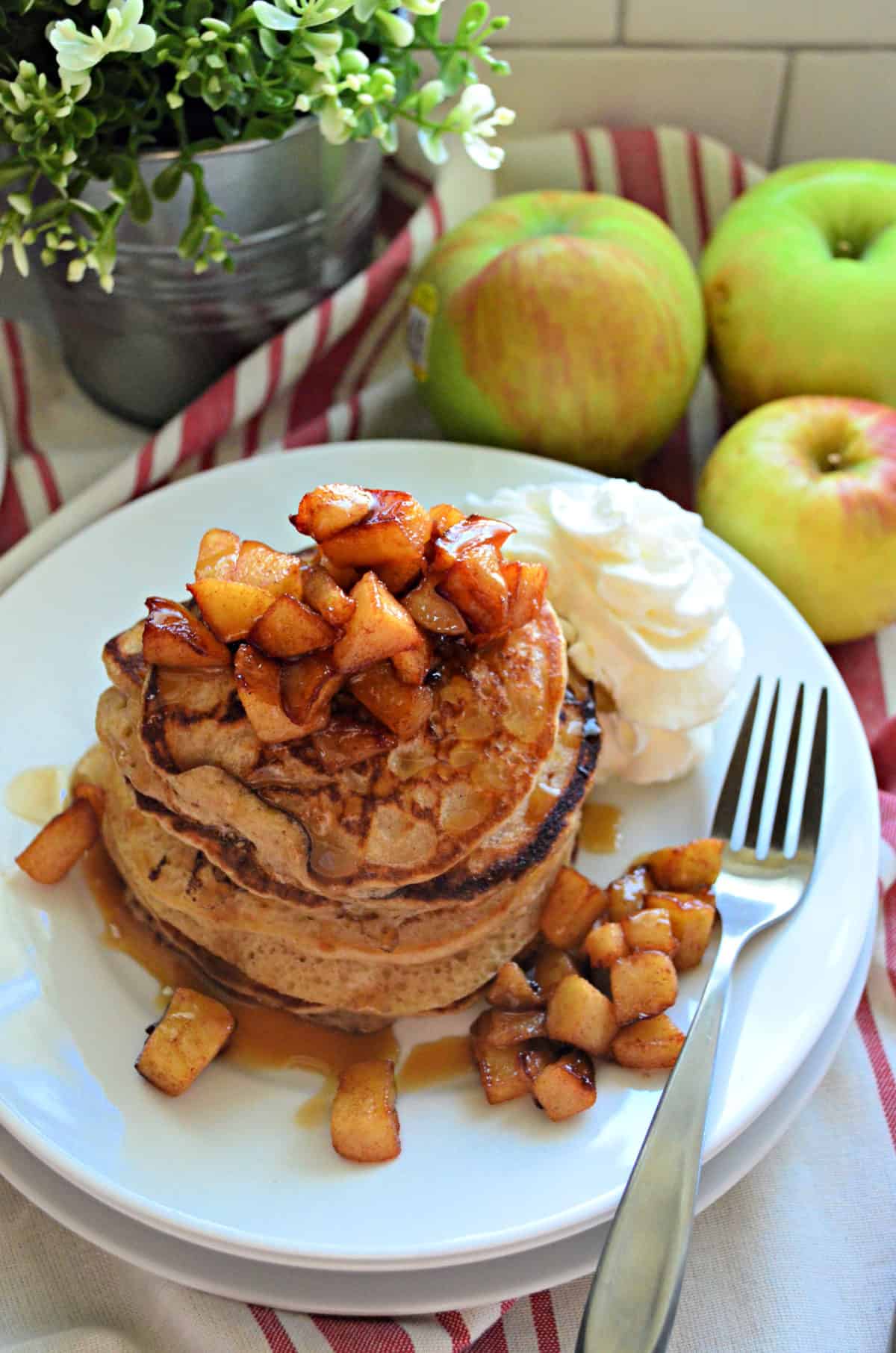 stack of cinnamon pancakes topped with cubed apples and syrup with whipped cream.