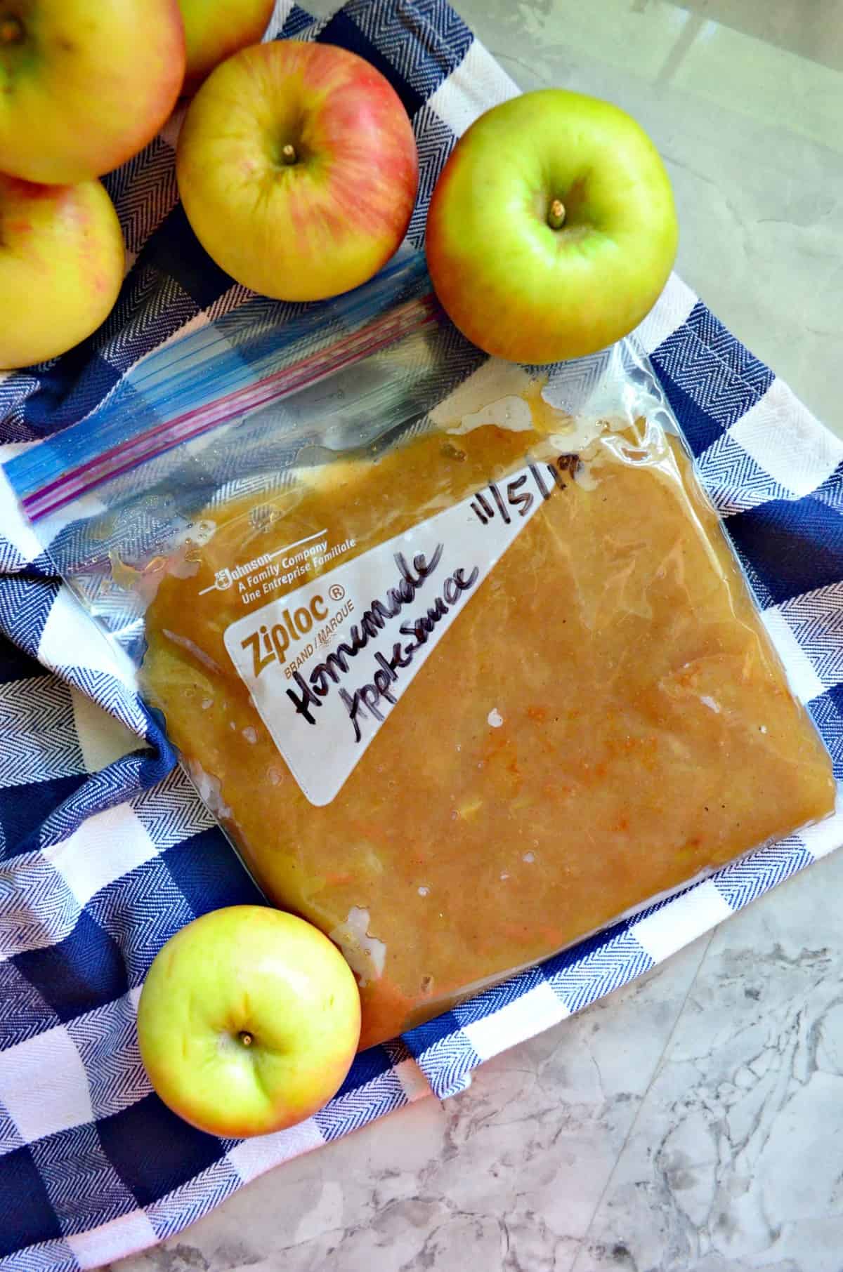 top view of labeled homemade applesauce in ziplock bag with apples on tablecloth.