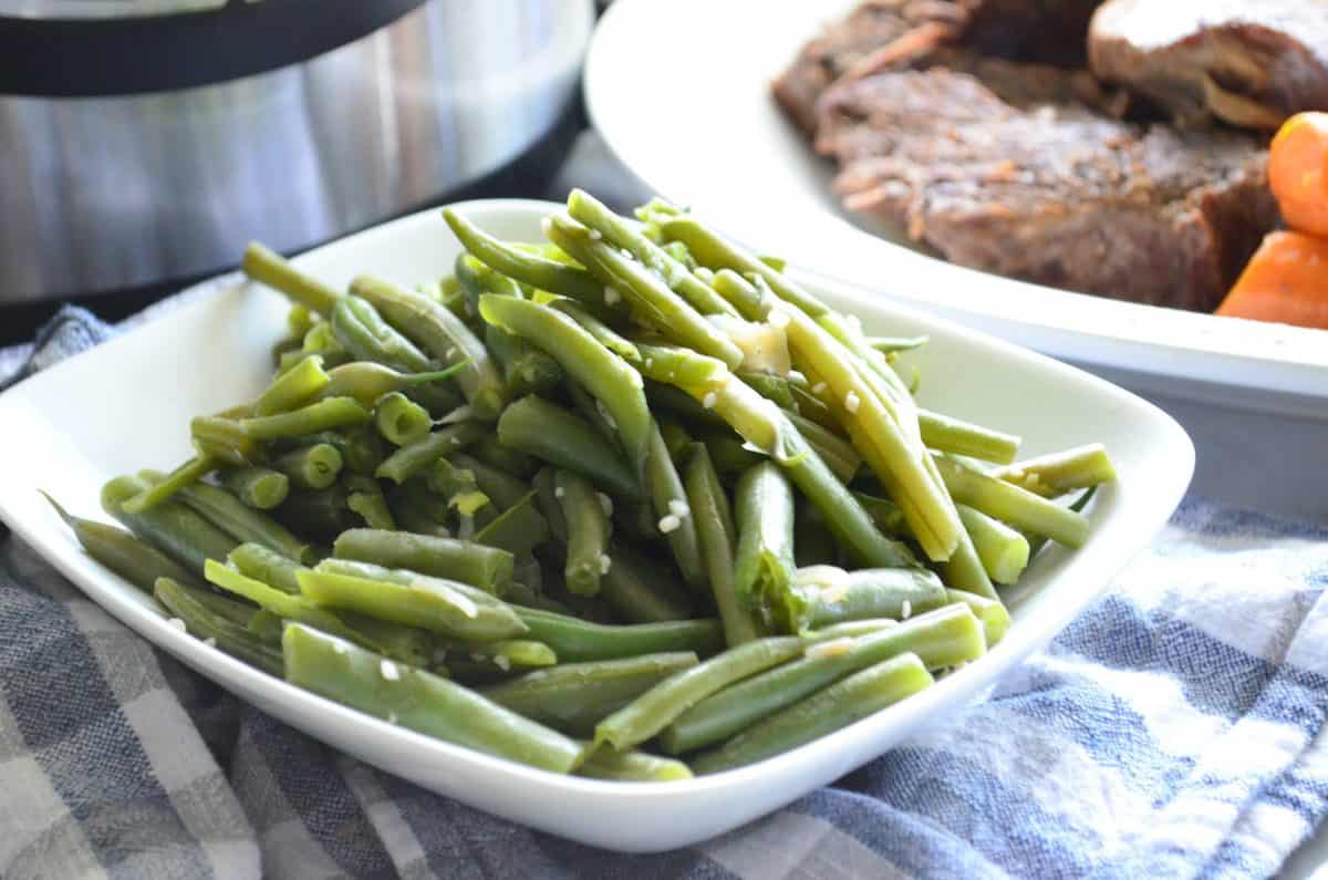 plated cooked green beans with minced garlic on top and plated pot roast in background.