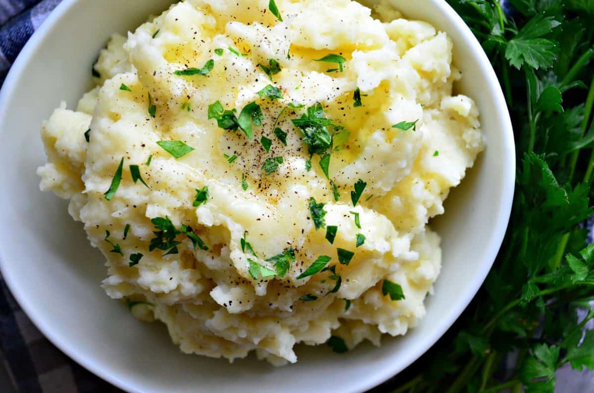 top view bowl of mashed potatoes topped with melted butter, pepper, and parsley next to parsley.