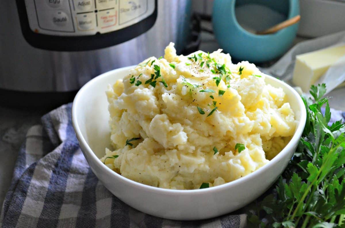 bowl of mashed potatoes topped with melted butter, pepper, and parsley on top of checkered tablecloth.