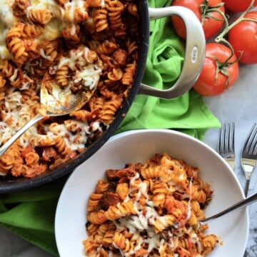 fusili with red sauce, melted cheese, and a spoon in a white bowl. with skillet next to it.