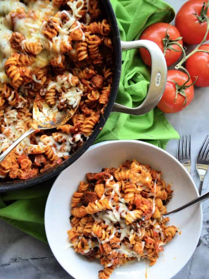 fusili with red sauce, melted cheese, and a spoon in a white bowl. with skillet next to it.