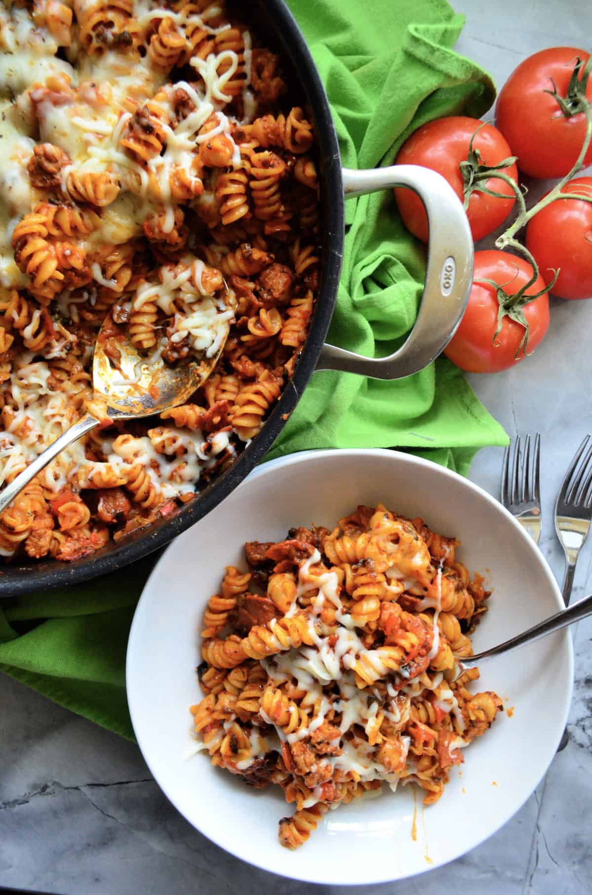 fusili with red sauce, melted cheese, and a spoon in a white bowl. with skillet next to it.