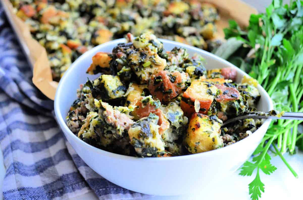 bowl full of stuffing with chopped green herbs in front of sheet pan with remnants and parsley.