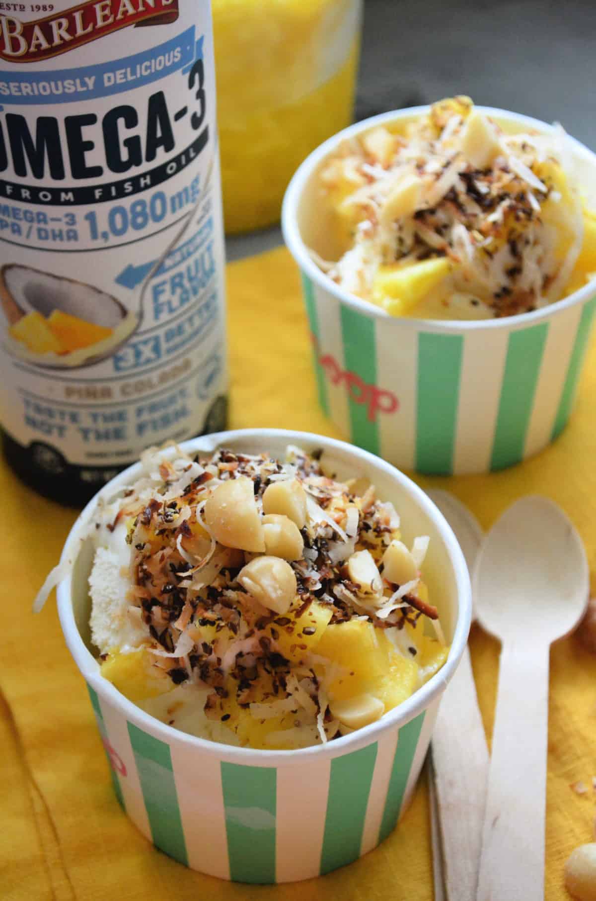 2 sundaes in paper cups topped with macadamia nuts, coconut shavings, and pineapple on yellow cloth.