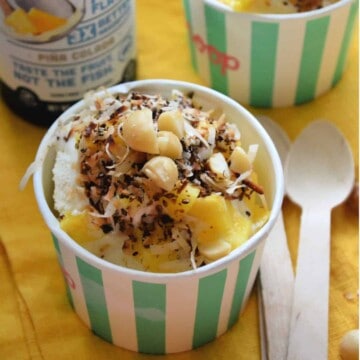 Closeup of frozen treat in paper cup topped with macadamia nuts, coconut shavings, and pineapple.