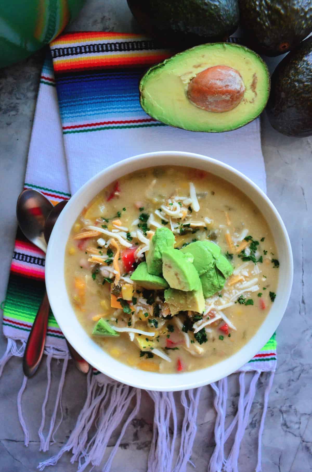 Green Enchilada Chicken Soup in bowl topped with cheese and avocado next to halve avocado.