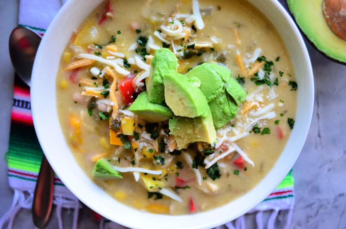 top view Green Enchilada Chicken Soup in bowl topped with cheese and avocado on colorful cloth.