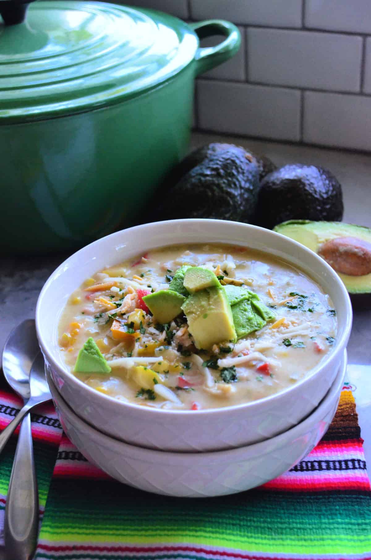 Bowl of green enchilada chicken soup stacked on top of empty bowl on colorful tablecloth with spoons.