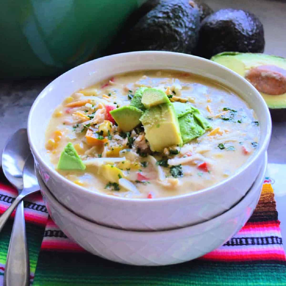 Two white bowls stacked with soup inside and chunks of avocado on top resting on a bright pink and green striped cloth.