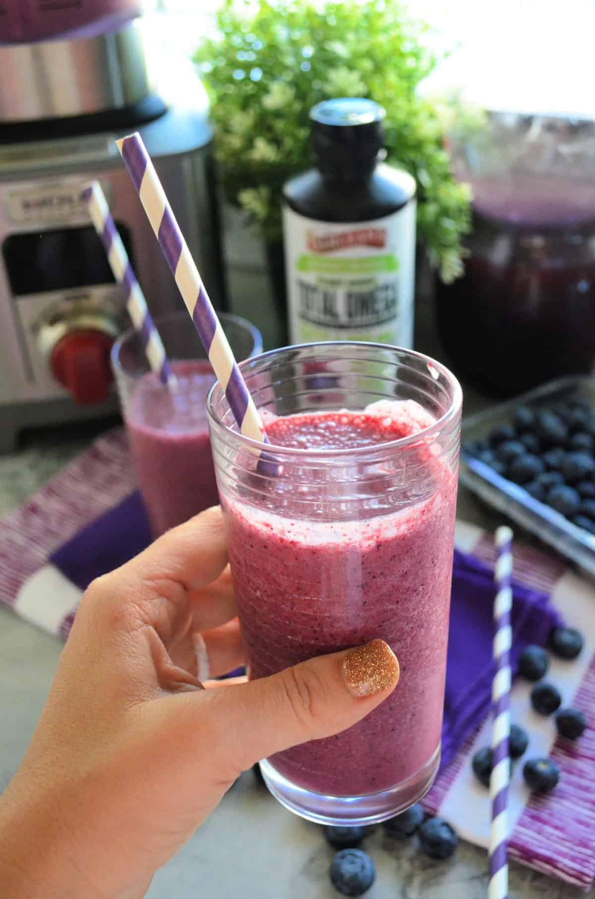 Hand holding up glass of purple smoothie with paper straw with smoothie in background.