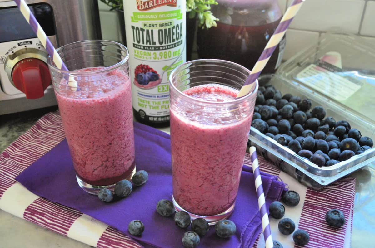 2 purple blended smoothies with paper straws on purple tablecloth with blueberries and total omega.