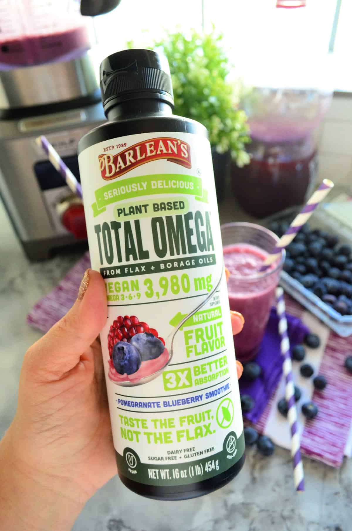 Barlean's Plant Based Total Omega from flax bottle being held close up with smoothies in background.