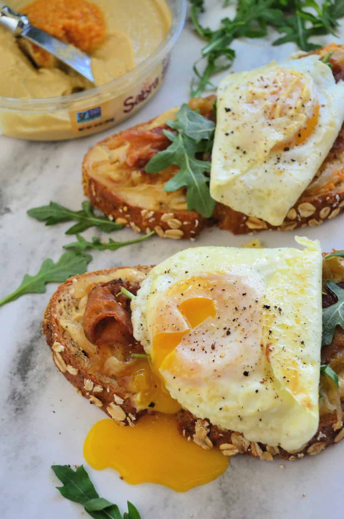 bread topped with hummus, bacon, over easy eggs oozing yolk, and arugula on platter.